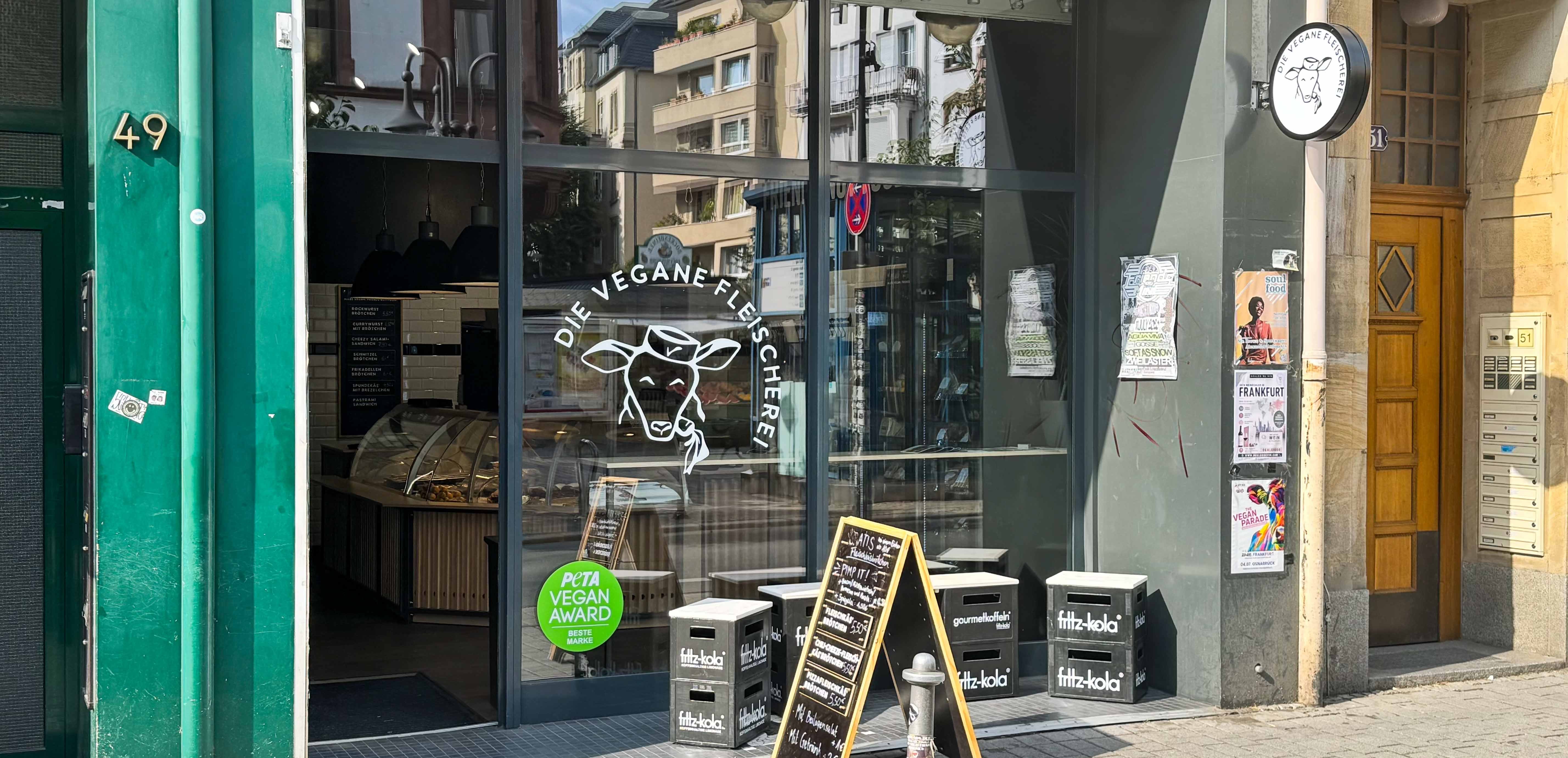Fassade von Die Vegane Fleischerei mit grünen Türen, Schaufenster mit Logo und veganen Produkten, PETA-Award-Schild und Fritz-Kola-Kisten draußen.