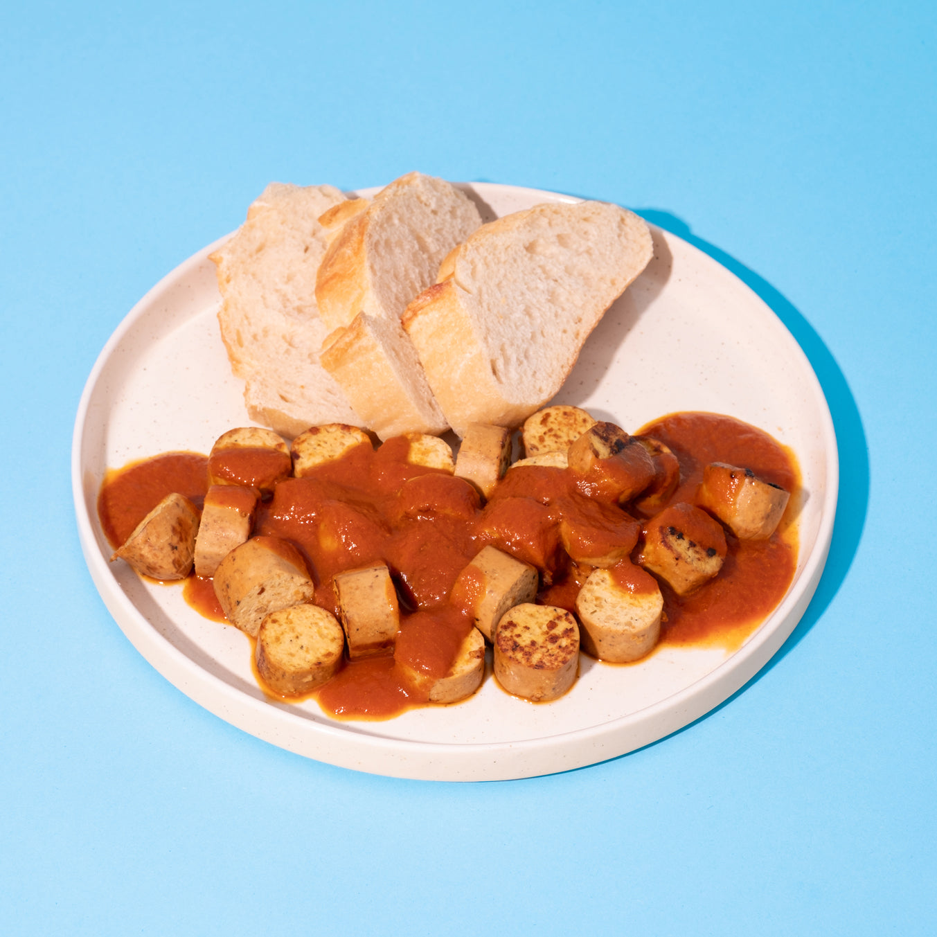 Vegane Currywurst mit Sauce und Brotscheiben auf weißem Teller vor blauem Hintergrund.