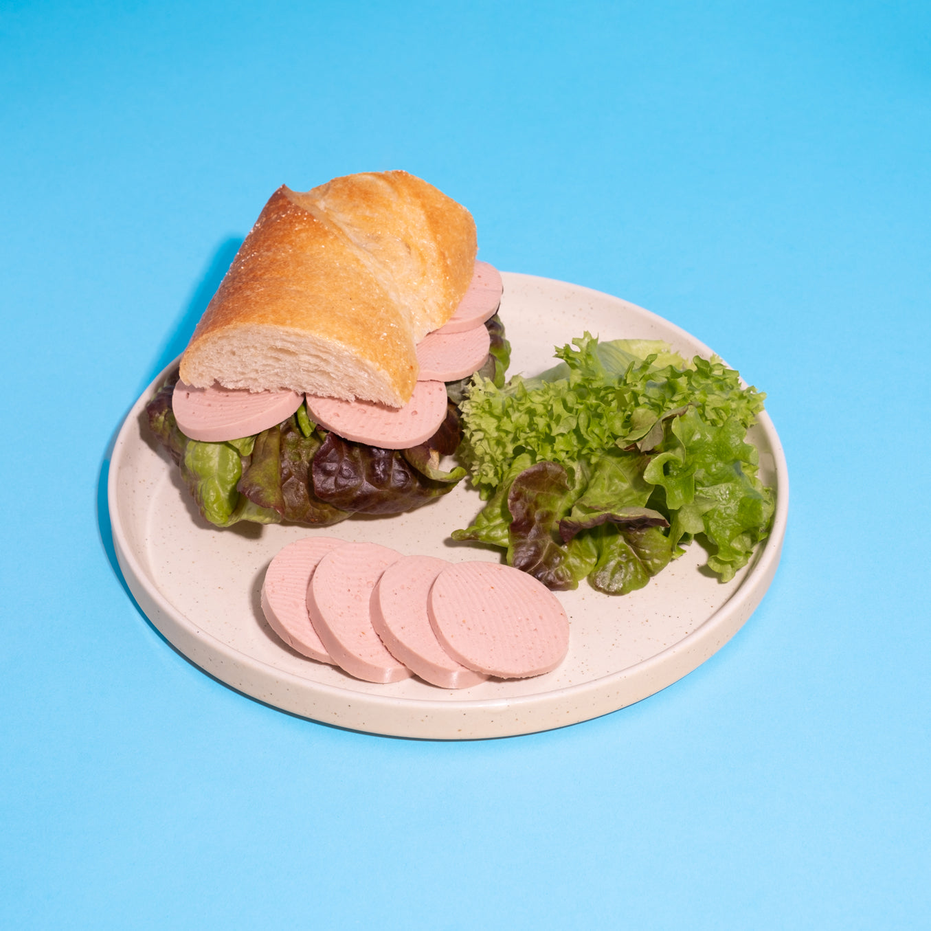 Veganes Lyoner-Natur-Sandwich mit Salat auf weißem Teller vor blauem Hintergrund.