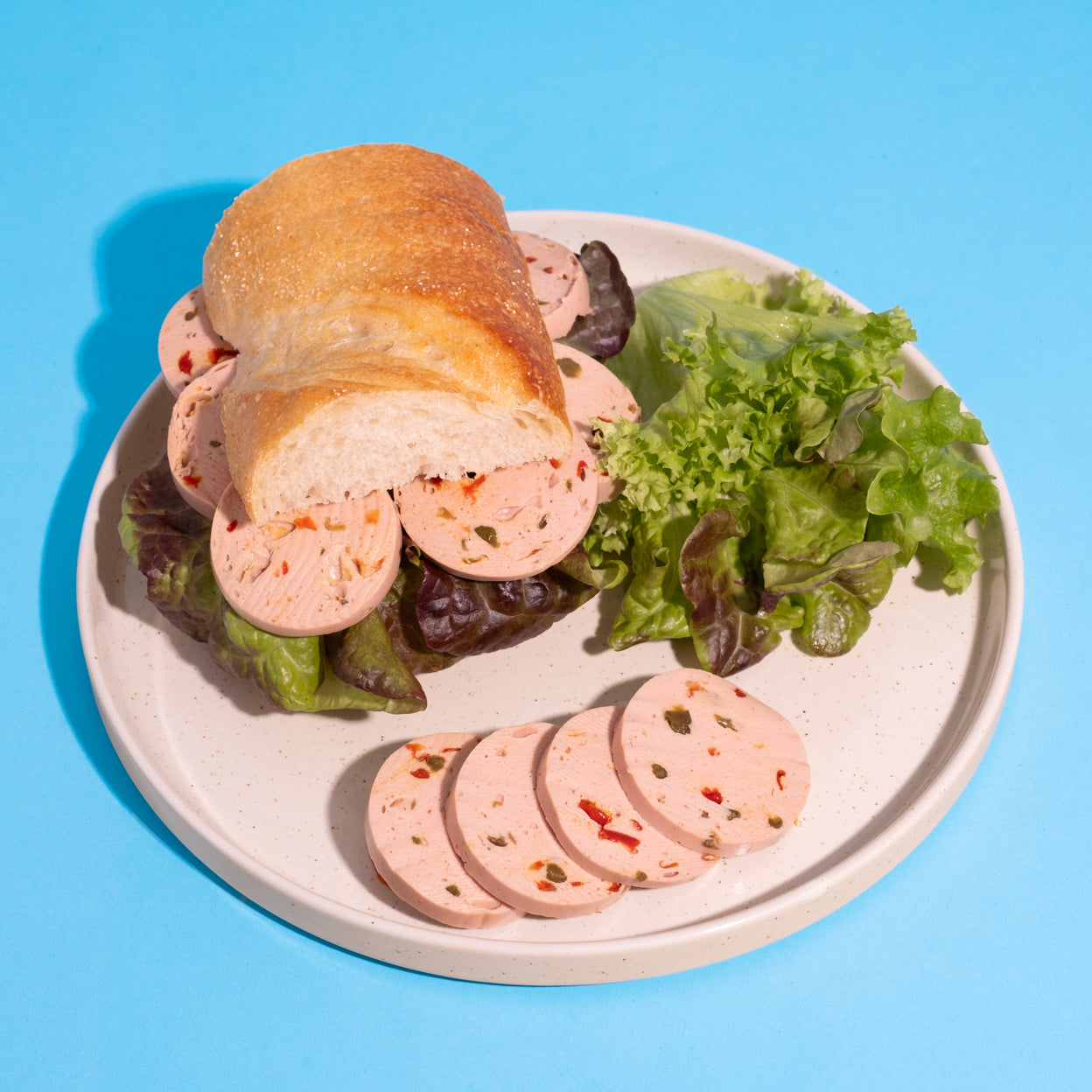 Veganes Lyoner-Paprika-Sandwich mit Salat auf weißem Teller vor blauem Hintergrund.