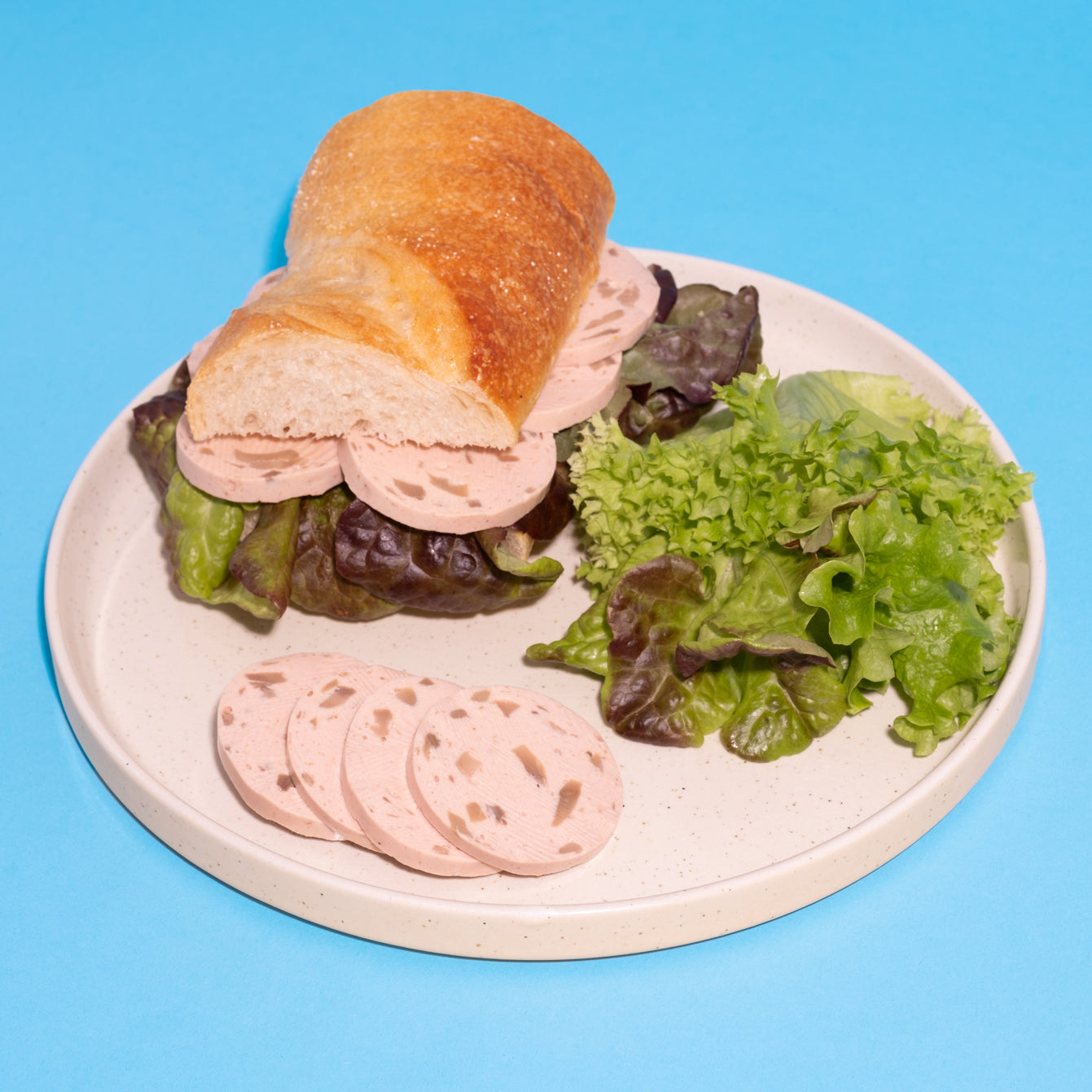 Veganes Lyoner-Pilz-Sandwich mit Salat auf weißem Teller vor blauem Hintergrund.