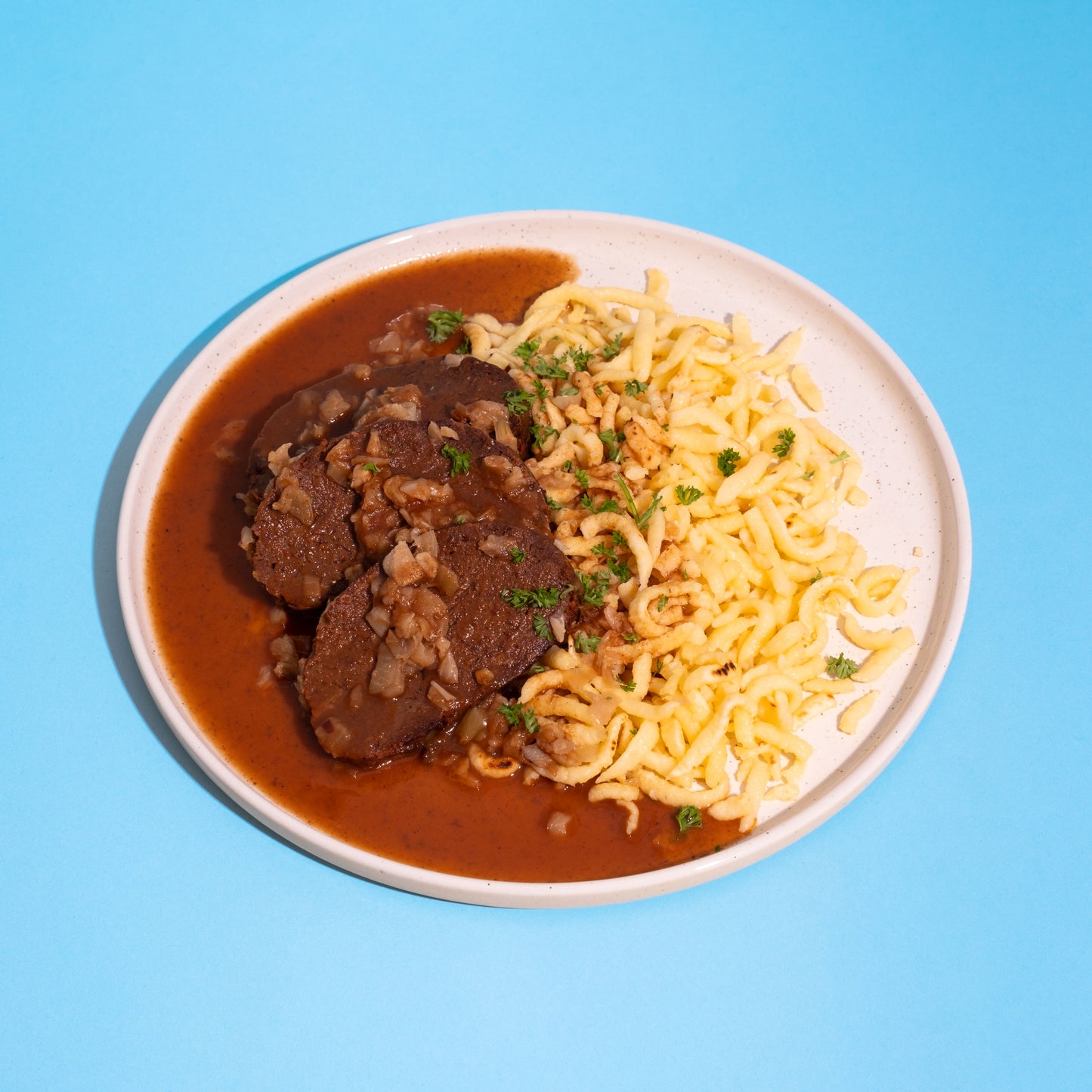 Teller mit zwei dunklen Seitanbraten in reichhaltiger brauner Soße, daneben goldgelbe Spätzle, garniert mit Petersilie auf.