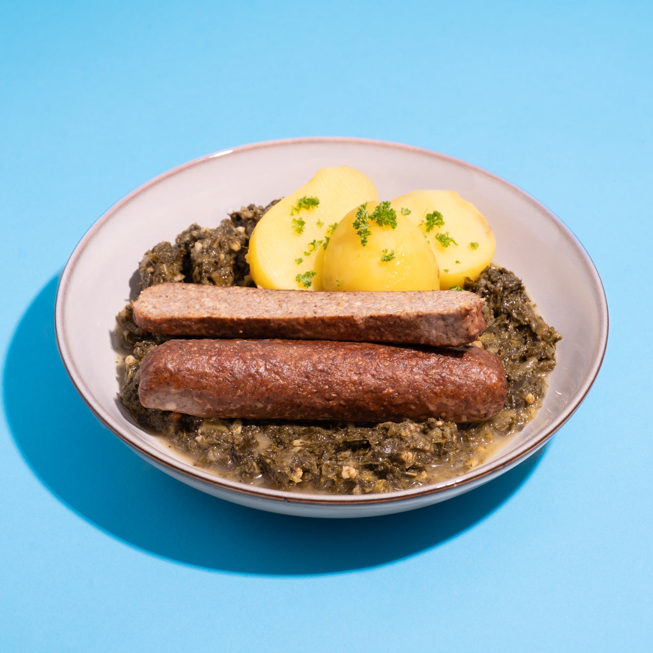 Teller mit veganer Pinkelwurst, Grünkohl und Kartoffeln vor blauem Hintergrund.