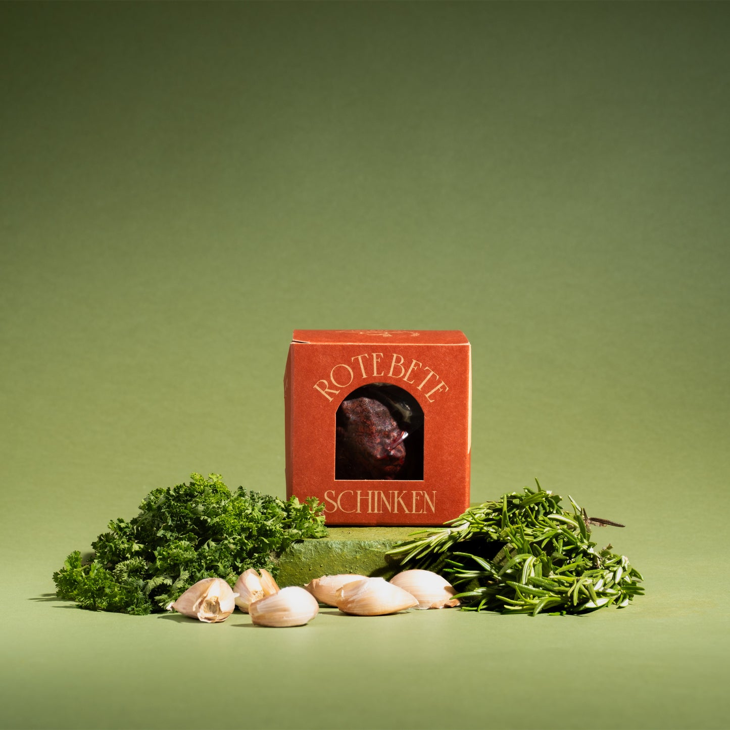 Veganes Rote Bete Schinken in roter Verpackung, umgeben von frischem Rosmarin, Petersilie und Knoblauchzehen – proteinreich & 100% pflanzlich