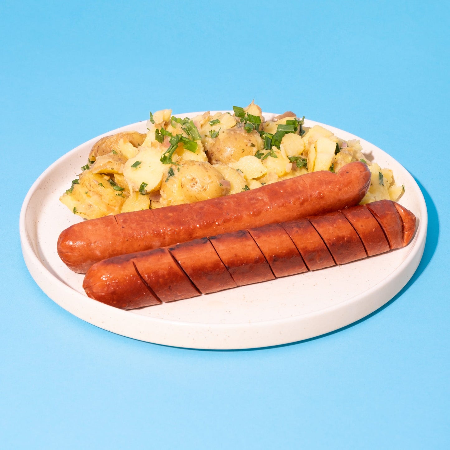 Vegane Bockwurst mit Kartoffelsalat auf weißem Teller vor blauem Hintergrund.