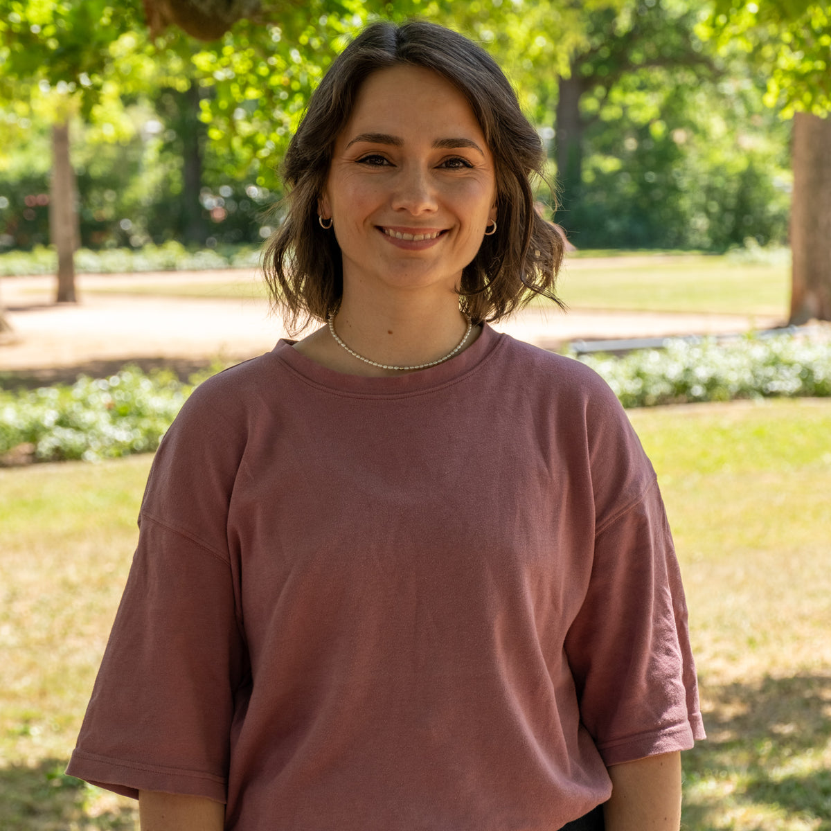Anika, Projektmanagement, lächelnd im Park in rosafarbenem T-Shirt mit Perlenkette