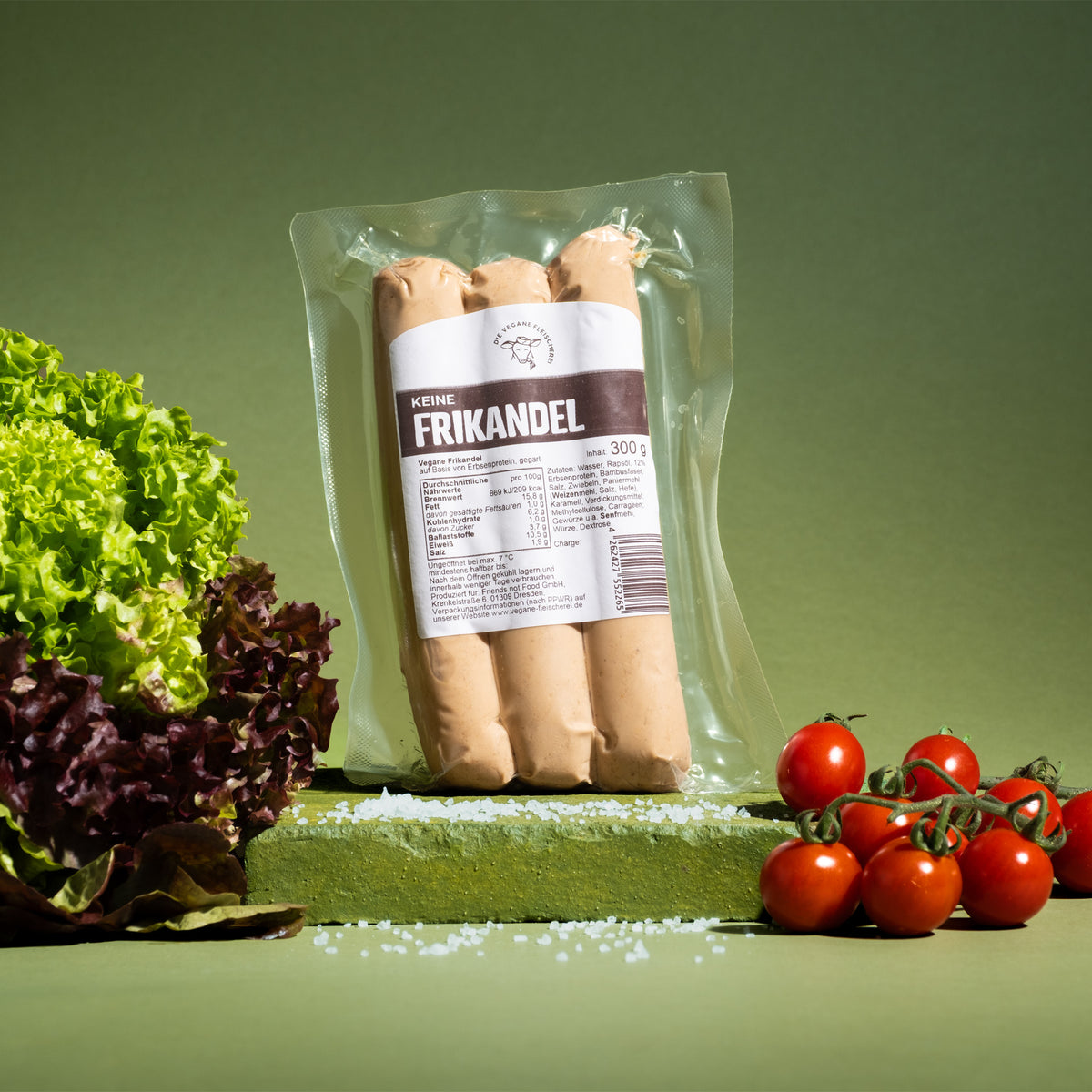 Packung „Keine Frikandel“, 300 g, mit drei Würsten, auf grünem Sockel mit Salat, Meersalz und Kirschtomaten