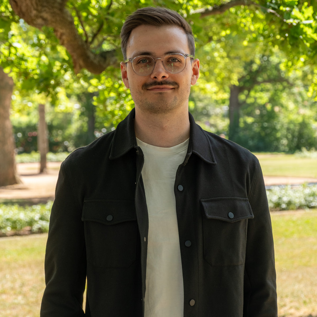 Jonathan, Leitung Onlineshop, im Park mit Brille und schwarzer Hemdjacke
