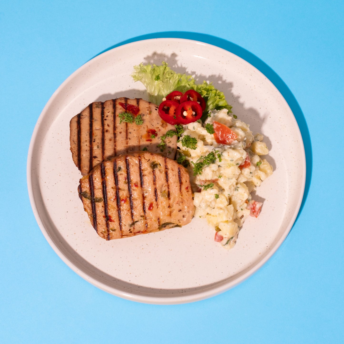 Zwei gegrillte Leberkäsescheiben mit Grillstreifen, dazu Kartoffelsalat mit Karottenstücken und Salatblätter auf weißem.