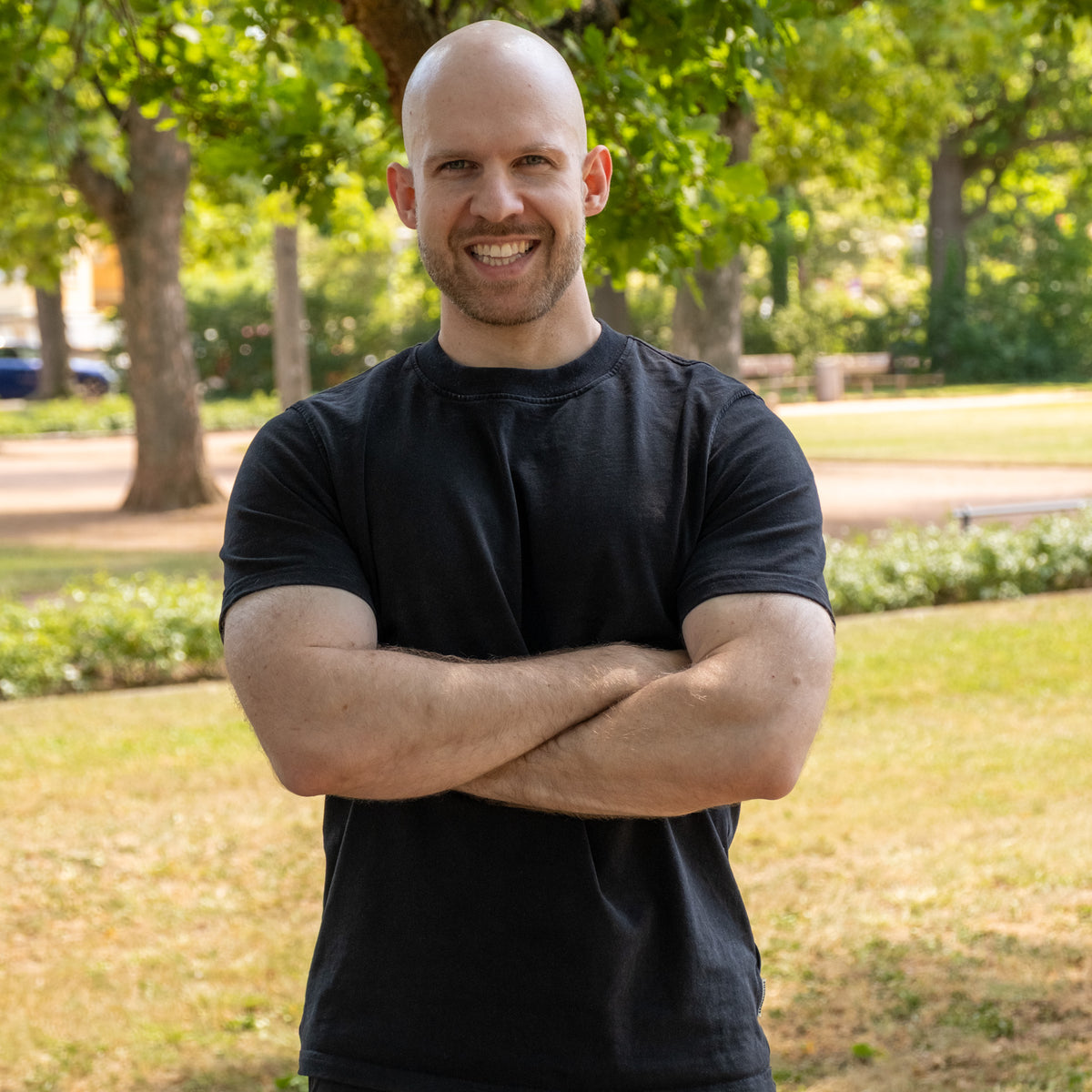 Nils, Geschäftsführer, lachend mit verschränkten Armen im Park, in schwarzem T-Shirt
