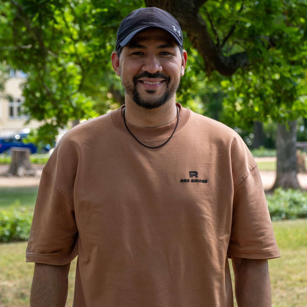 Ridouane aus der Produktion, lächelnd im Park mit schwarzer Kappe und braunem T-Shirt