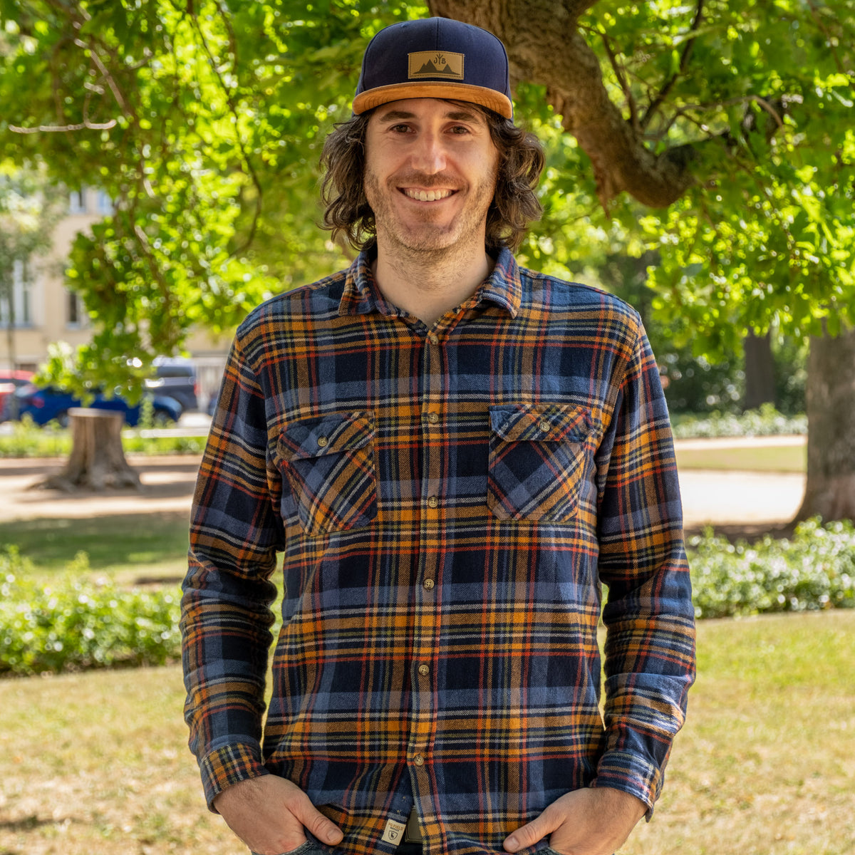 Stefan aus der Produktentwicklung, lächelnd im Park mit Kappe und kariertem Hemd