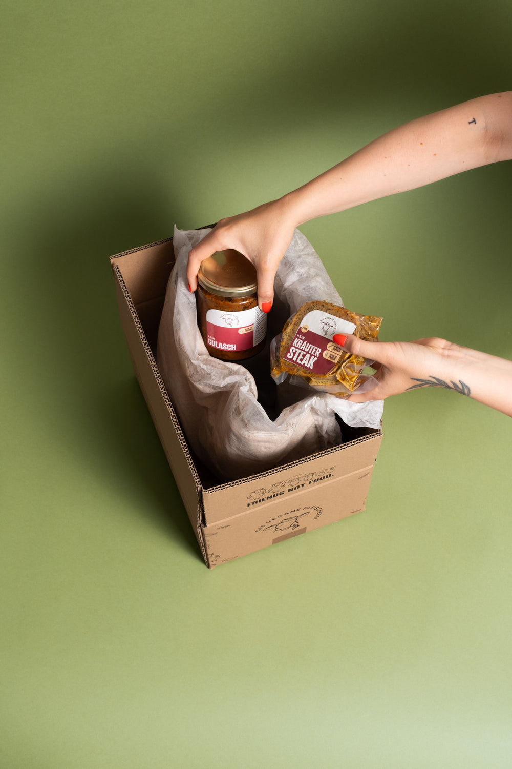 Hände packen ein Glas veganes Gulasch und ein vakuumiertes „Kein Kräutersteak“ in einen mit Papier ausgeschlagenen Versandkarton
