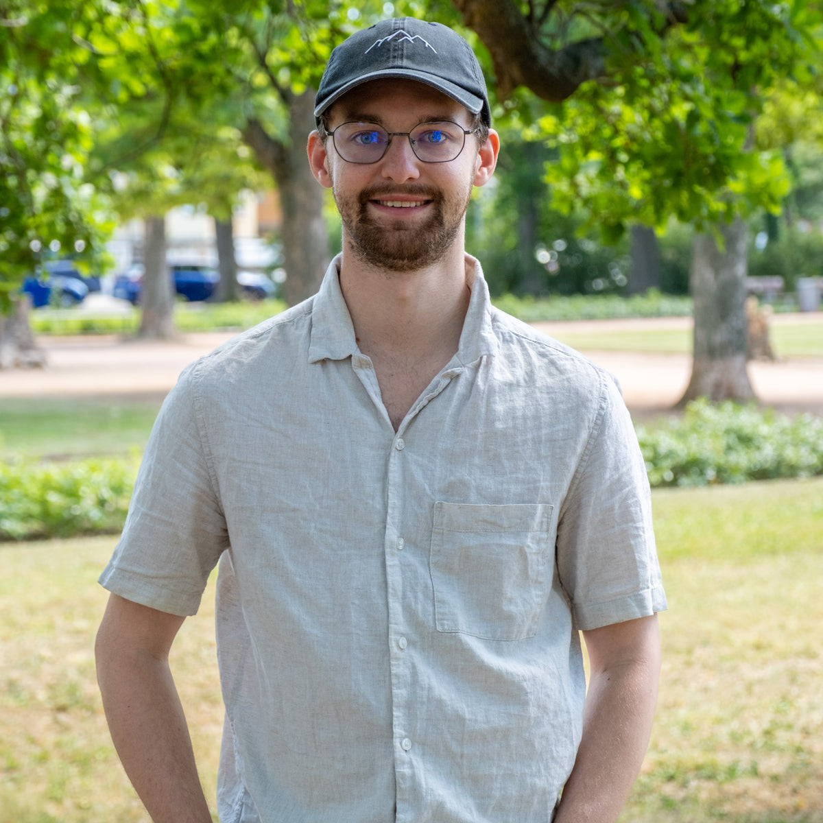 Leo, Leitung Produktion, lächelnd im Park mit Kappe, Brille und hellem Leinenhemd
