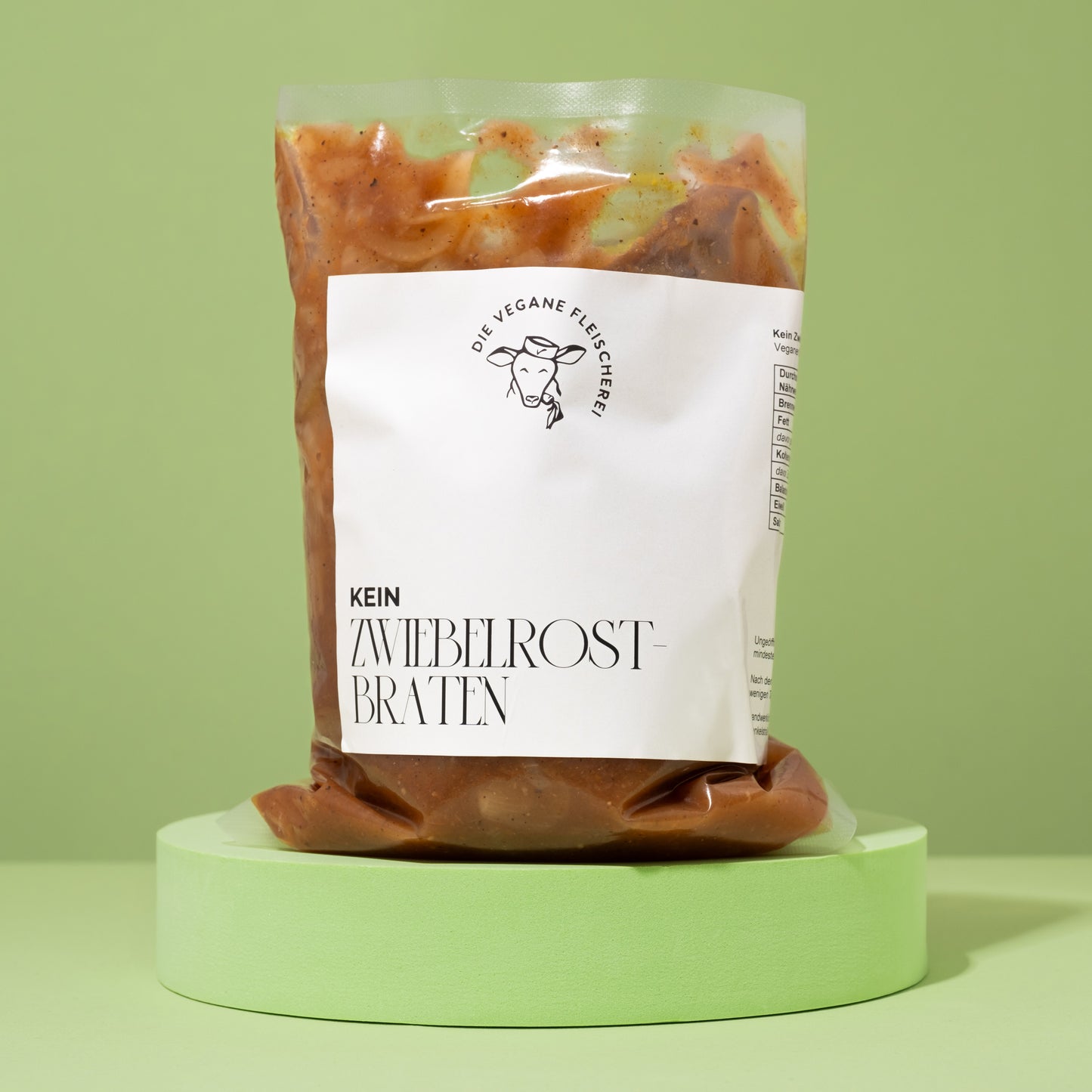 Vakuumverpackter, brauner Seitan-Zwiebelrostbraten mit Soße, auf rundem, pastellgrünem Podest vor grünem Hintergrund.