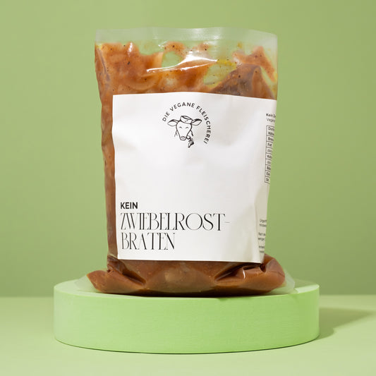 Vakuumverpackter, brauner Seitan-Zwiebelrostbraten mit Soße, auf rundem, pastellgrünem Podest vor grünem Hintergrund.