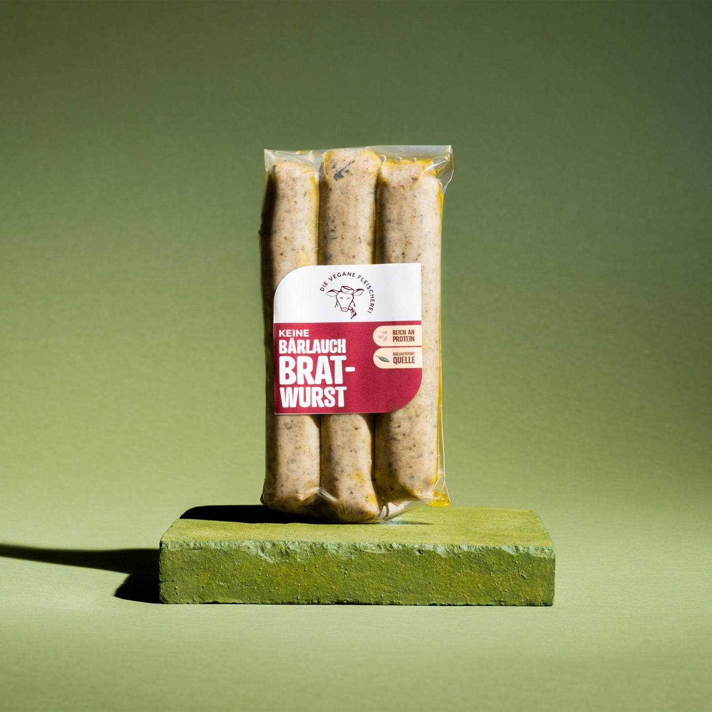 Vegane Bratwurst, proteinreich & pflanzlich, in transparenter Verpackung auf grünem Podest
