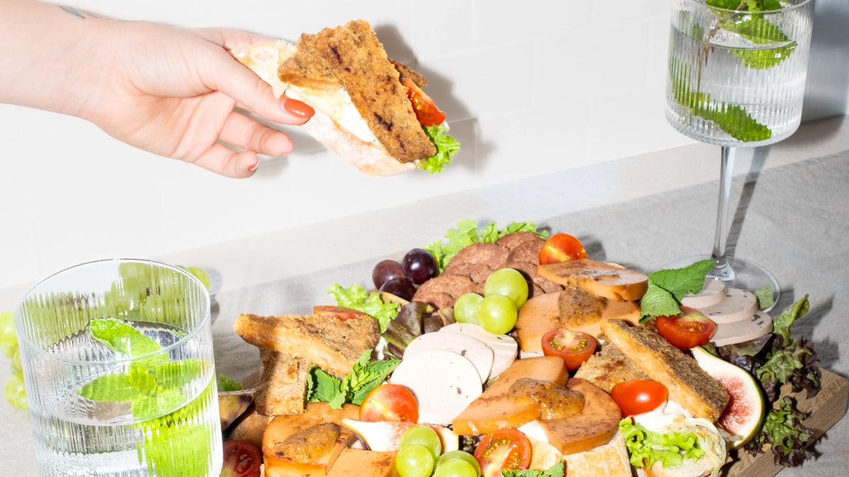 Hand hält ein belegtes Sandwich über eine Imbissplatte mit veganem Aufschnitt, Trauben und Tomaten