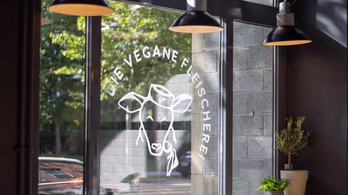 Schaufenster einer Filiale mit weißem Logo „Die Vegane Fleischerei“ mit Kuhkopf, dahinter Hängelampen und Bäume