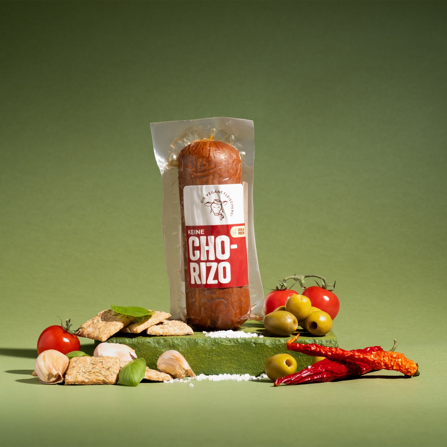Vegane Chorizo-Wurst in Verpackung, umgeben von Tomaten, Oliven, Knoblauch und Chili auf grünem Podest