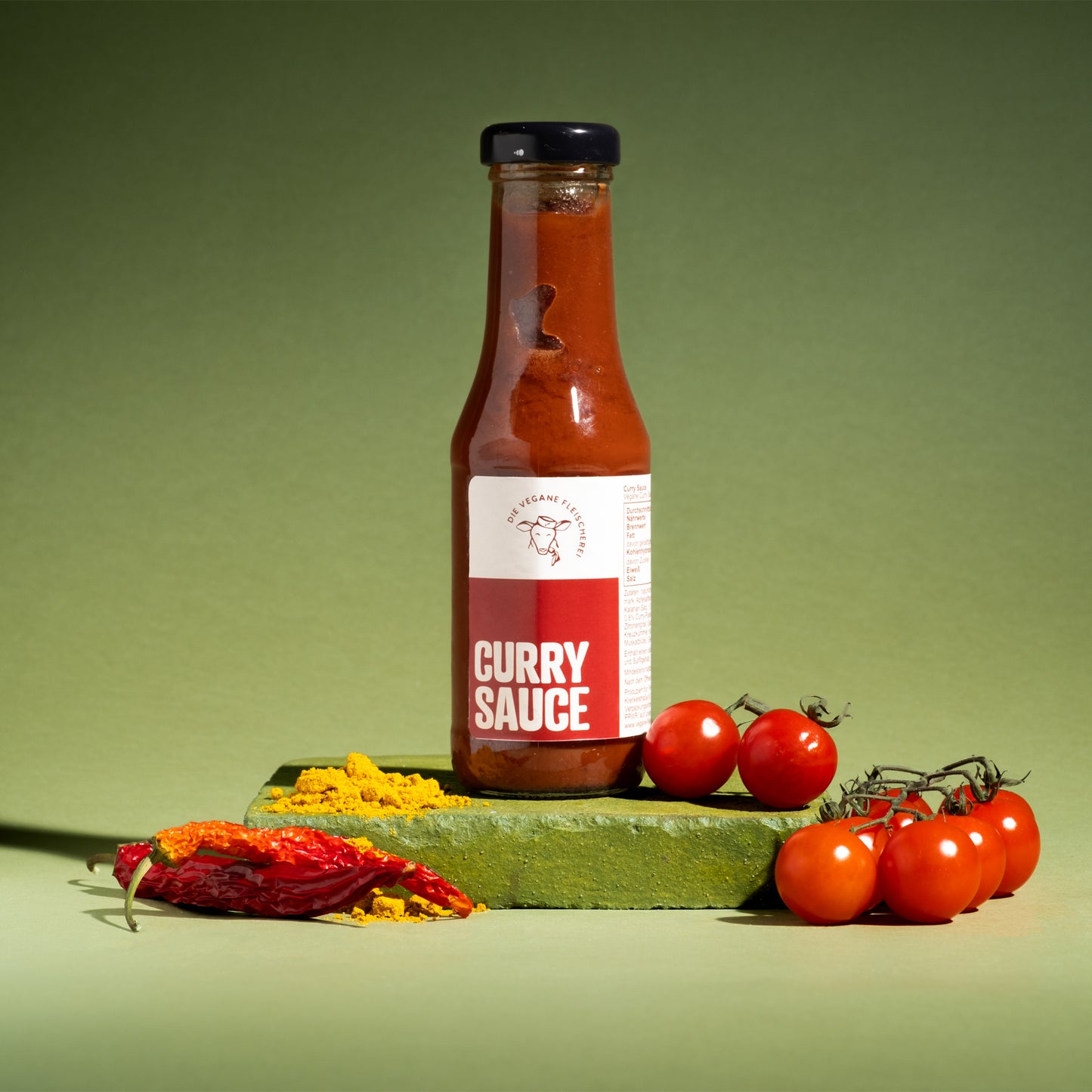Vegane Curry Sauce in Glasflasche mit frischen Tomaten, roten Chilis und Kurkuma auf grünem Stein