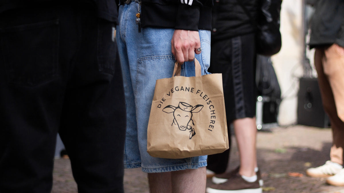 Person hält eine Papiertüte mit Logo und Schriftzug „Die Vegane Fleischerei“ in der Hand