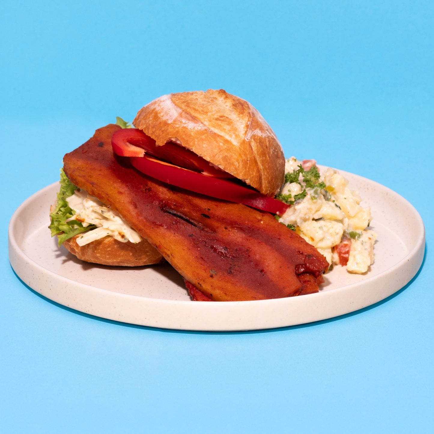 Veganer Grill-Bacon im Brötchen mit Kartoffelsalat auf weißem Teller.