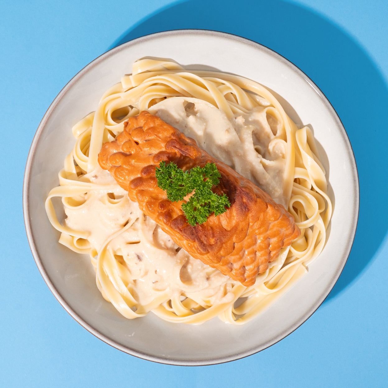 Veganes Laxfilet mit Pasta und heller Sauce in weißer Schüssel vor blauem Hintergrund. 