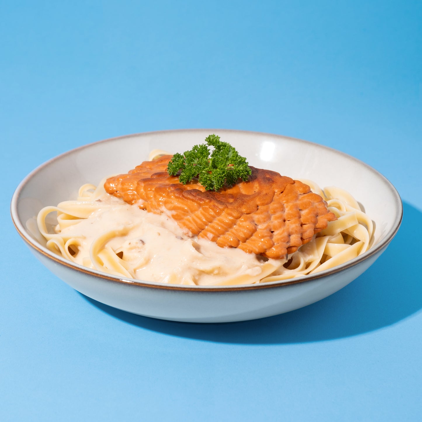 Veganes Laxfilet mit Pasta und heller Sauce in weißer Schüssel vor blauem Hintergrund.