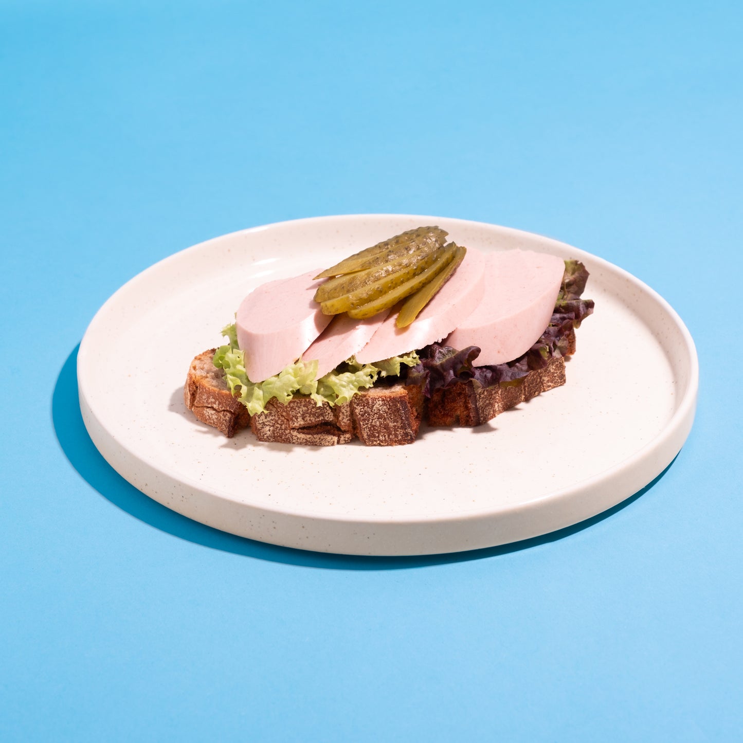 Belegtes Brot mit veganer Fleischwurst, Salat und Gewürzgurke auf weißem Teller vor blauem Hintergrund.
