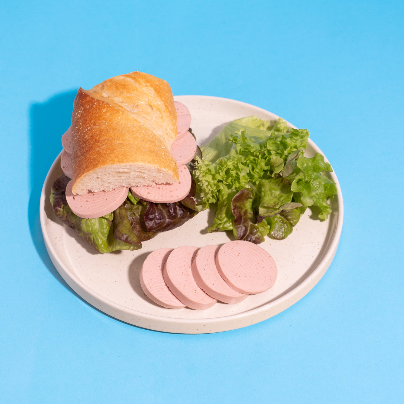 Veganes Lyoner-Natur-Sandwich mit Salat auf weißem Teller vor blauem Hintergrund. 