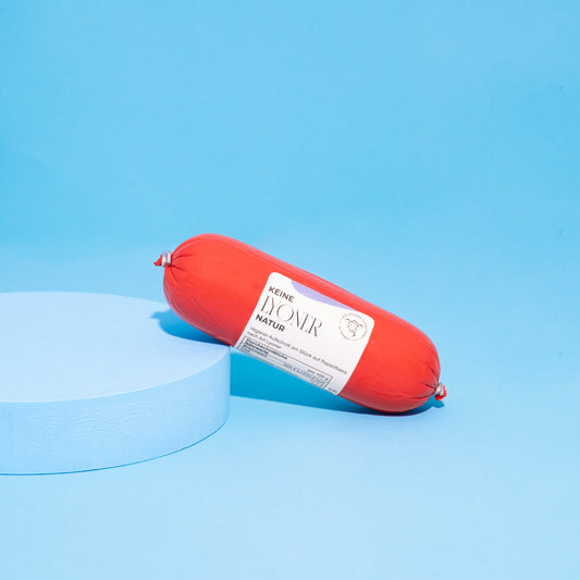 Vegane Lyoner Natur in Verpackung auf blauem Podest vor blauem Hintergrund. 