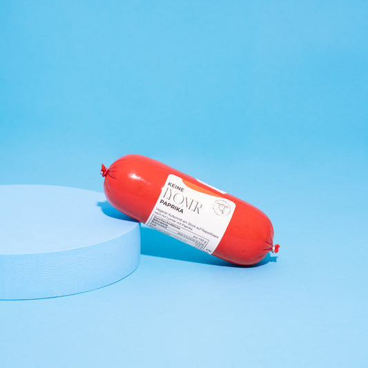 Vegane Lyoner Paprika in Verpackung auf blauem Podest vor blauem Hintergrund. 