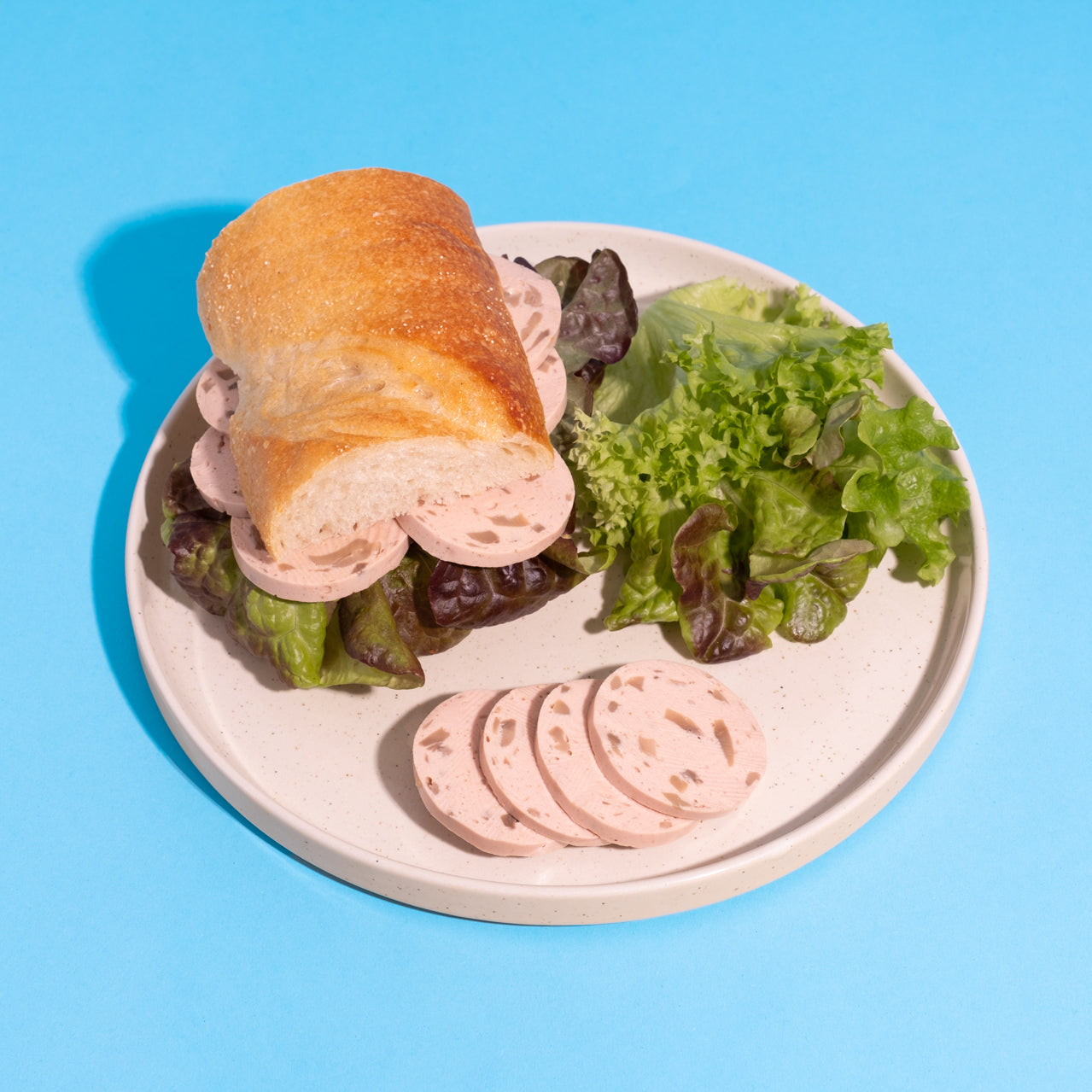 Veganes Lyoner-Pilz-Sandwich mit Salat auf weißem Teller vor blauem Hintergrund. 