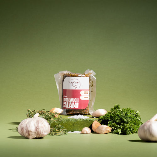 Verpackte „Keine Knoblauchsalami“ auf grünem Podest, umgeben von Knoblauchknollen, -zehen, Thymian, Petersilie und Salz