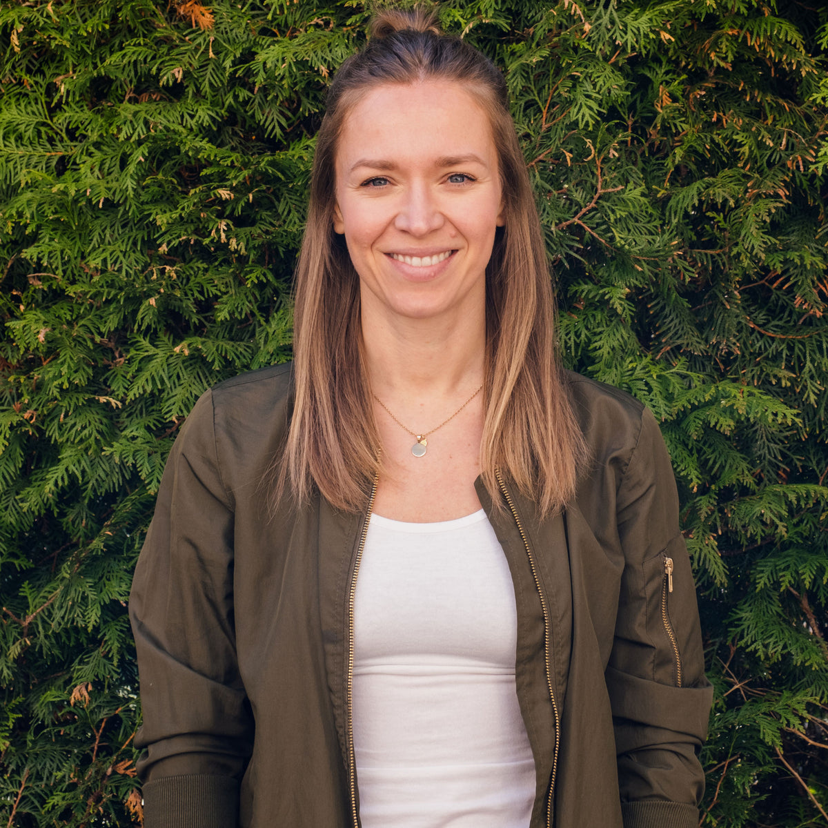 Marina aus der Teamassistenz, lächelnd in olivgrüner Jacke und weißem Shirt vor einer grünen Hecke