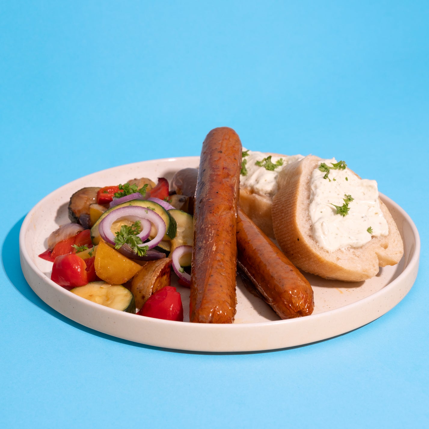 Teller mit veganer mediterraner Bratwurst und Gemüse vor blauem Hintergrund.