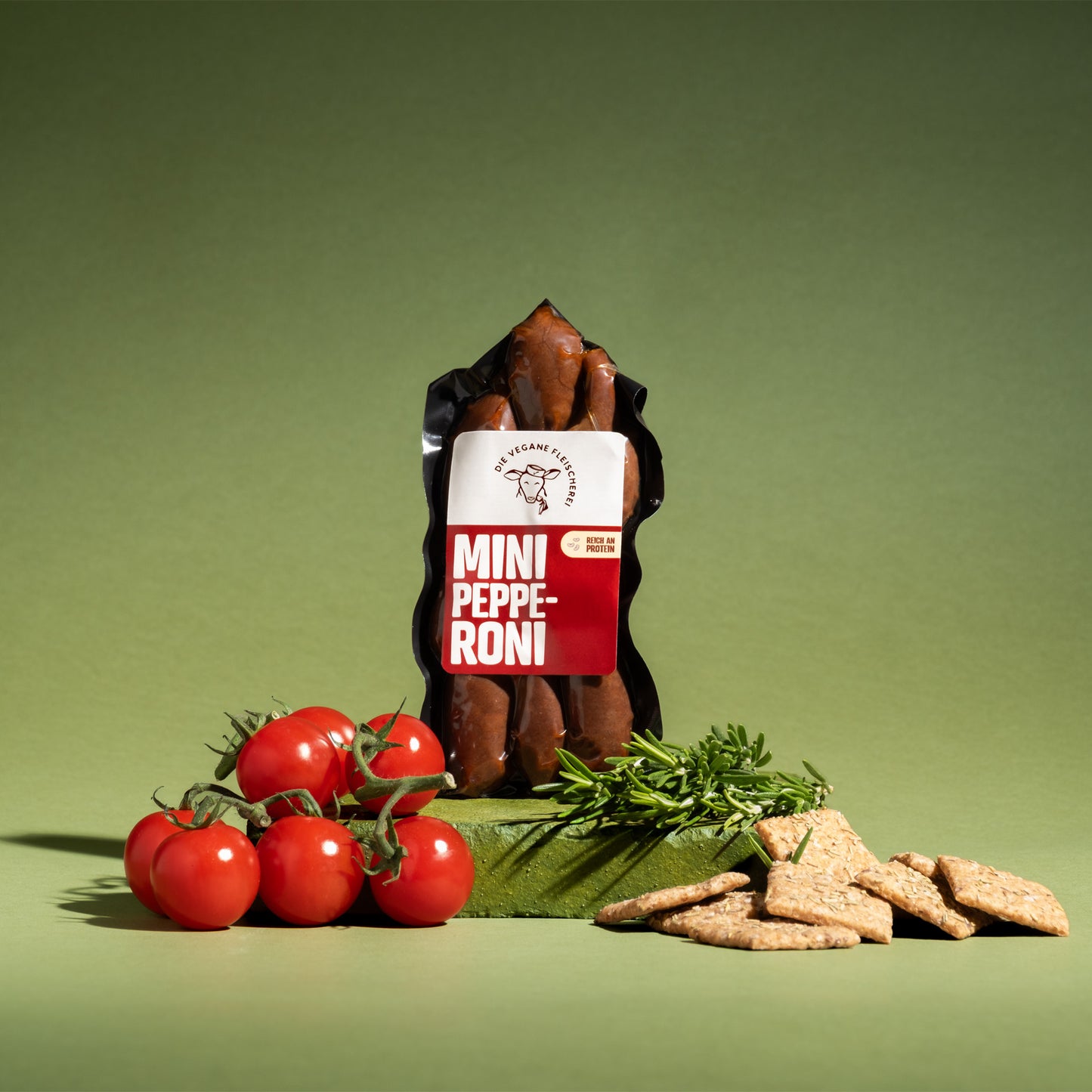 Vegane Mini Pepperoni, proteinreich, mit Kirschtomaten, Rosmarin und Crackern auf grünem Hintergrund