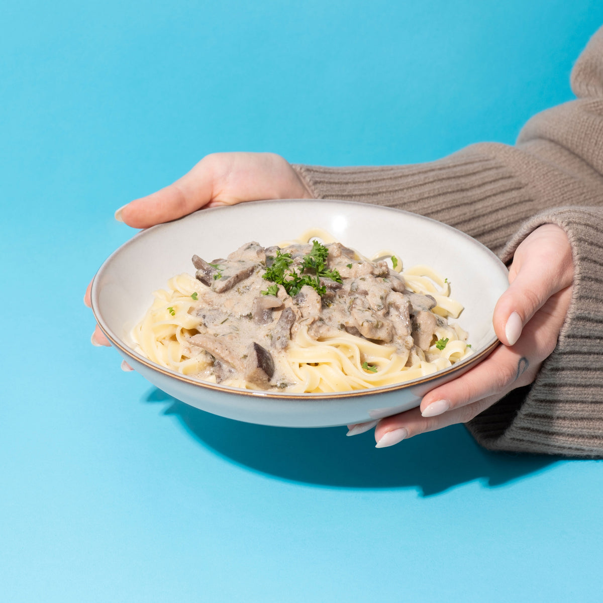 Zwei Hände in beigem Strickpullover halten Teller mit Tagliatelle und „Kein Pilzrahm-Geschnetzeltes“, garniert mit Petersilie