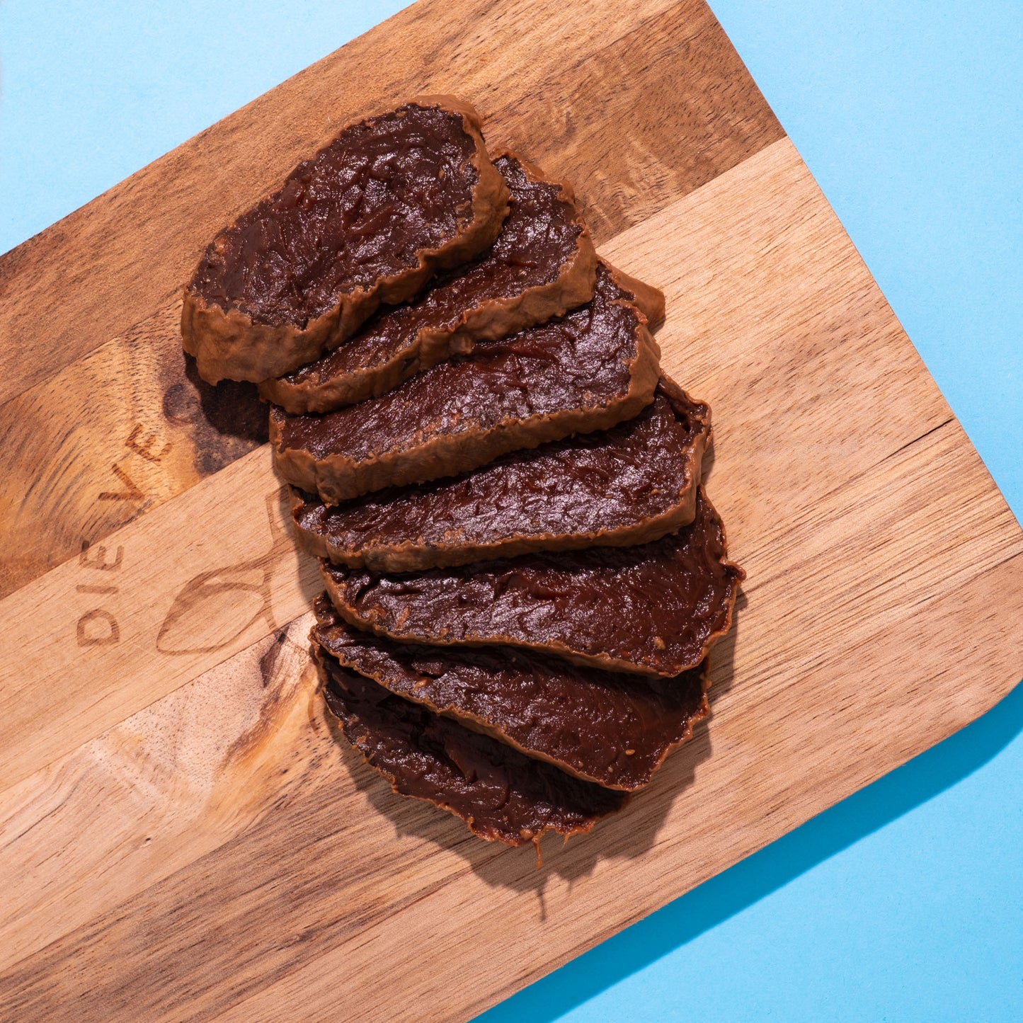 Mehrere dünne Scheiben dunkelroter Rote-Bete-Schinken auf hellem Holzbrett, blauer Hintergrund.
