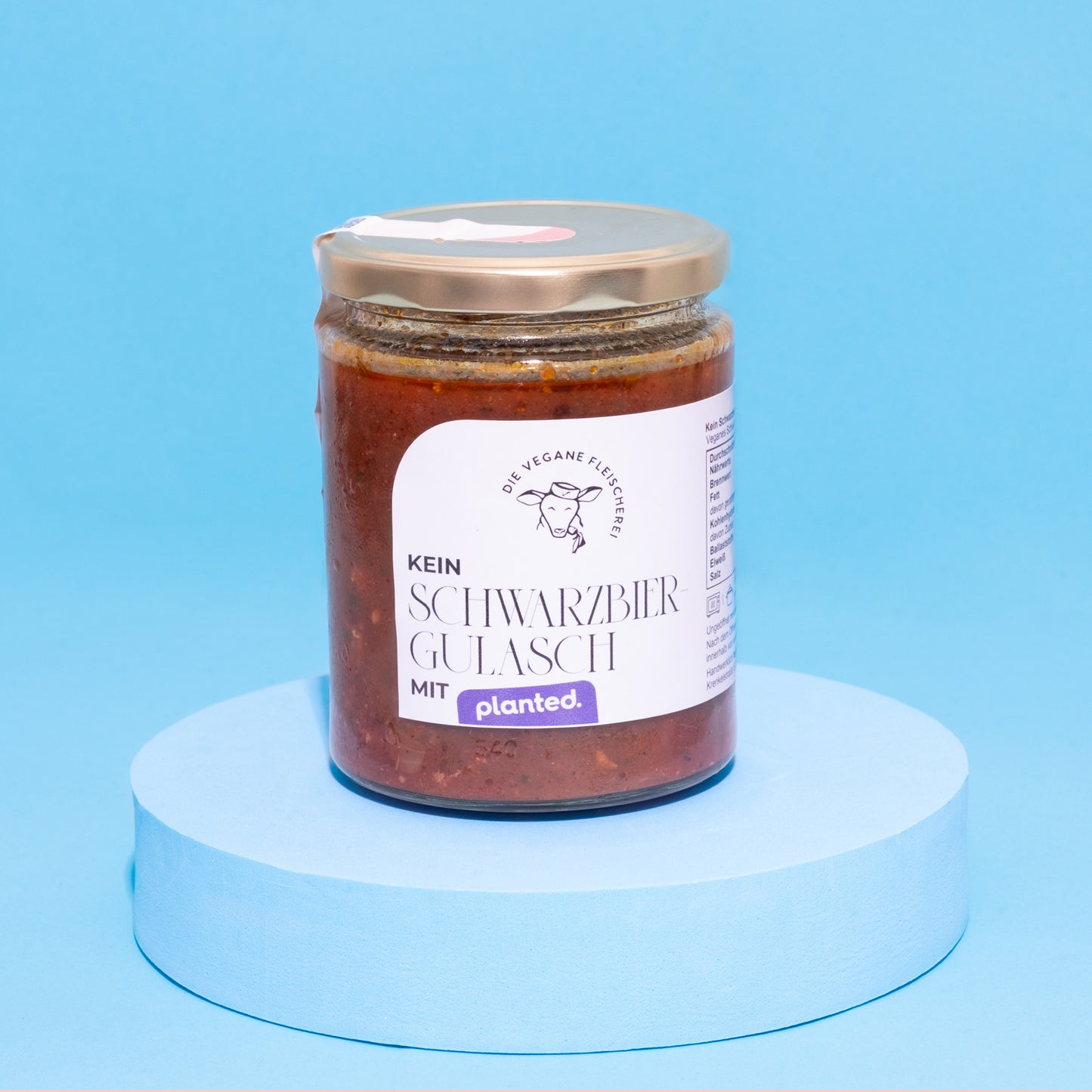 Glas mit veganem Schwarzbiergulasch auf blauem Podest vor blauem Hintergrund.