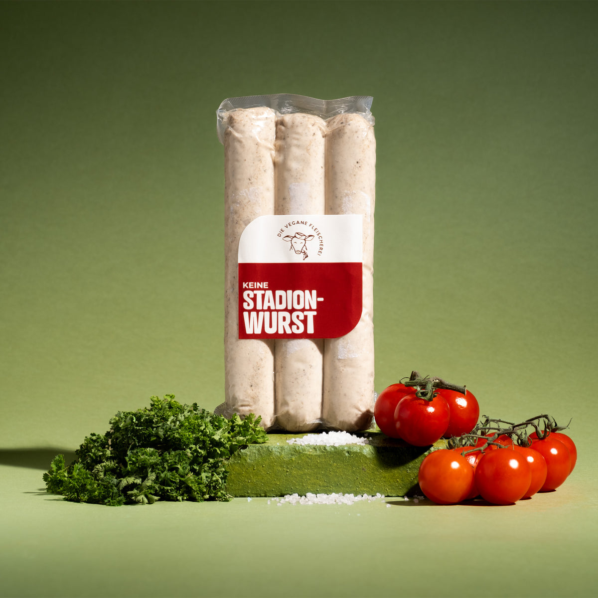 Drei Würste „Keine Stadionwurst“ in Vakuumpackung, daneben krause Petersilie, grobes Salz und Kirschtomaten