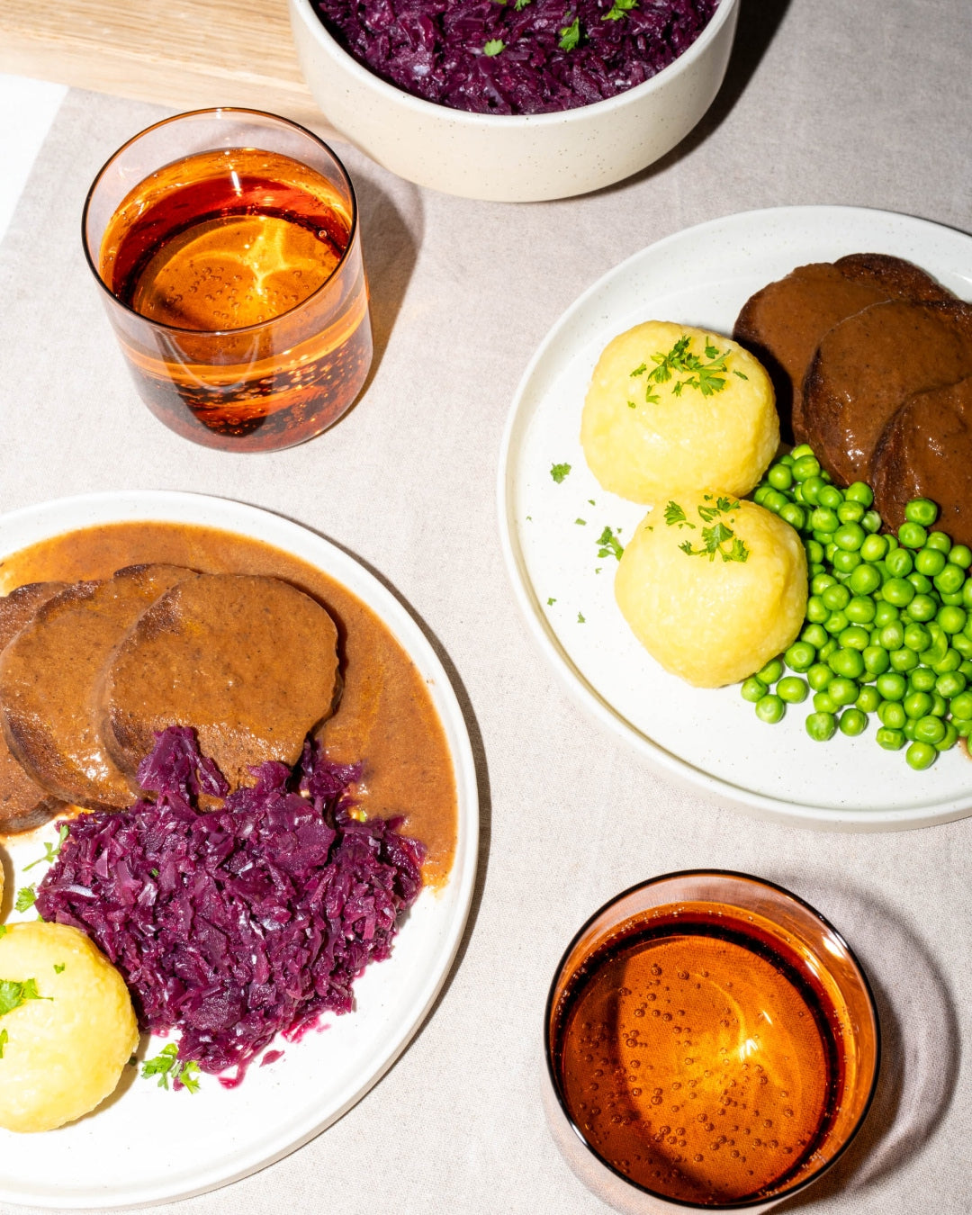 Zwei Teller mit veganem Braten in Soße, Rotkohl, Kartoffelklößen und Erbsen, dazu Gläser mit Getränken
