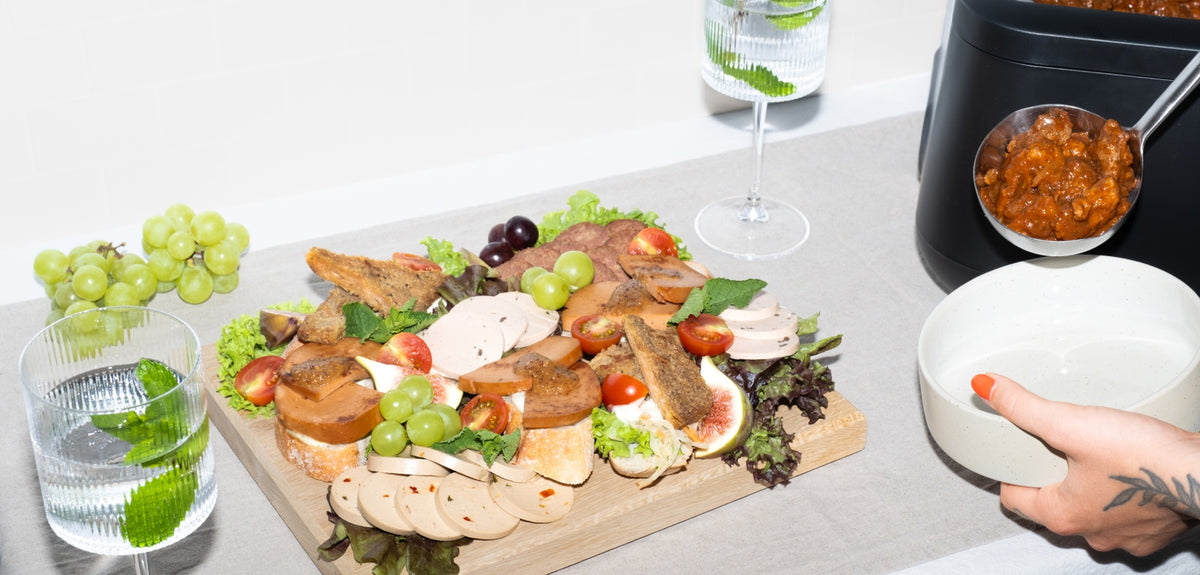 Catering-Platte mit veganem Aufschnitt, Weintrauben, Feigen und Tomaten auf einem Holzbrett, daneben wird Gulasch aus einem Topf in eine Schale geschöpft