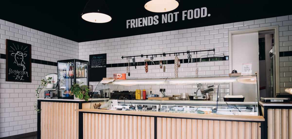 Filiale der Veganen Fleischerei mit weiß gefliesten Wänden, Schriftzug „Friends not Food.“, hängenden Würsten und holzverkleideter Theke