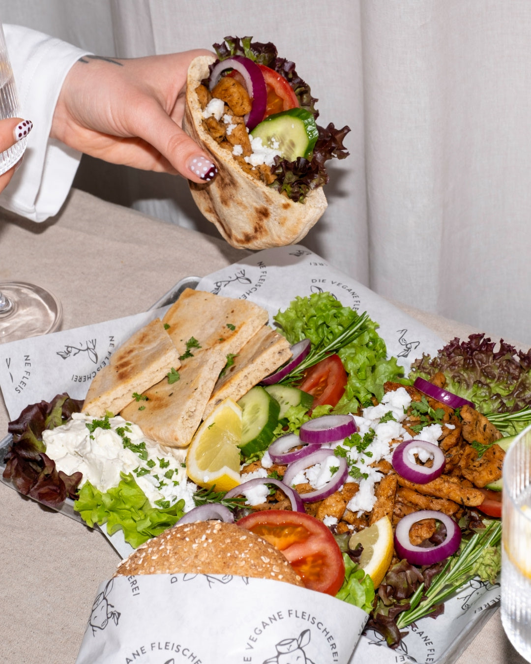 Gefülltes Fladenbrot mit angebratenen veganen Fleischstreifen, Salat und Zwiebeln über einem Tablett mit Fladenbrot, weißem Dip, Tomaten und Zitrone