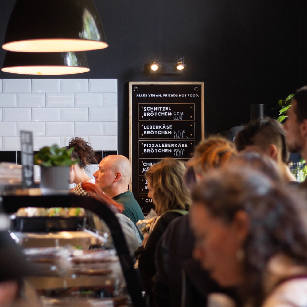 Gäste in einer Filiale vor einer Tafel mit „Alles vegan. Friends not Food.“ und Brötchenangebot wie Schnitzel- und Leberkäsebrötchen