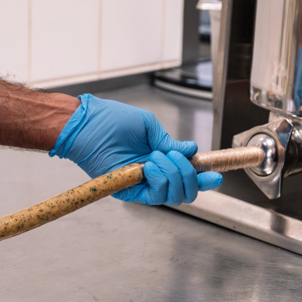 Hand in blauem Handschuh führt eine frisch gefüllte Wurst mit Kräutern aus der Füllmaschine in der Manufaktur